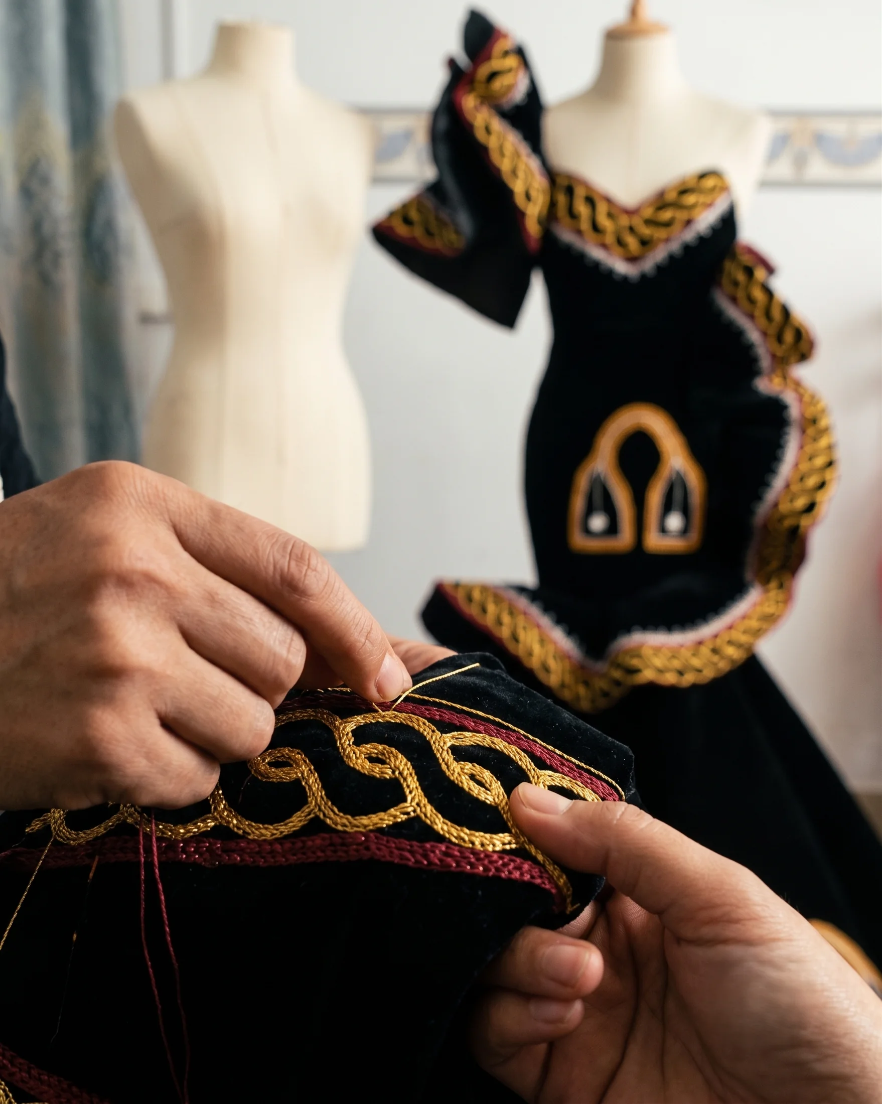 Hands sewing gold and red embroidery onto a black gown